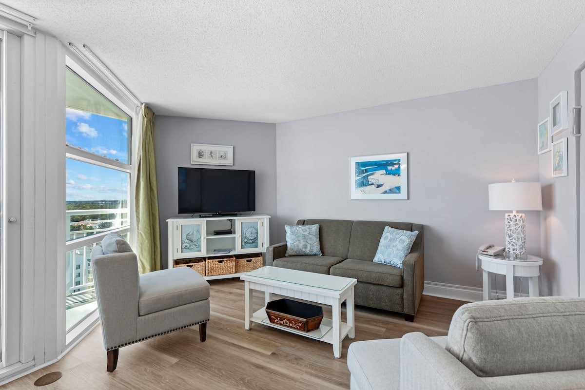 A modern living room with a grey sofa, a white coffee table, and a television on a white cabinet. There are wicker baskets on the floor and a window with curtains.