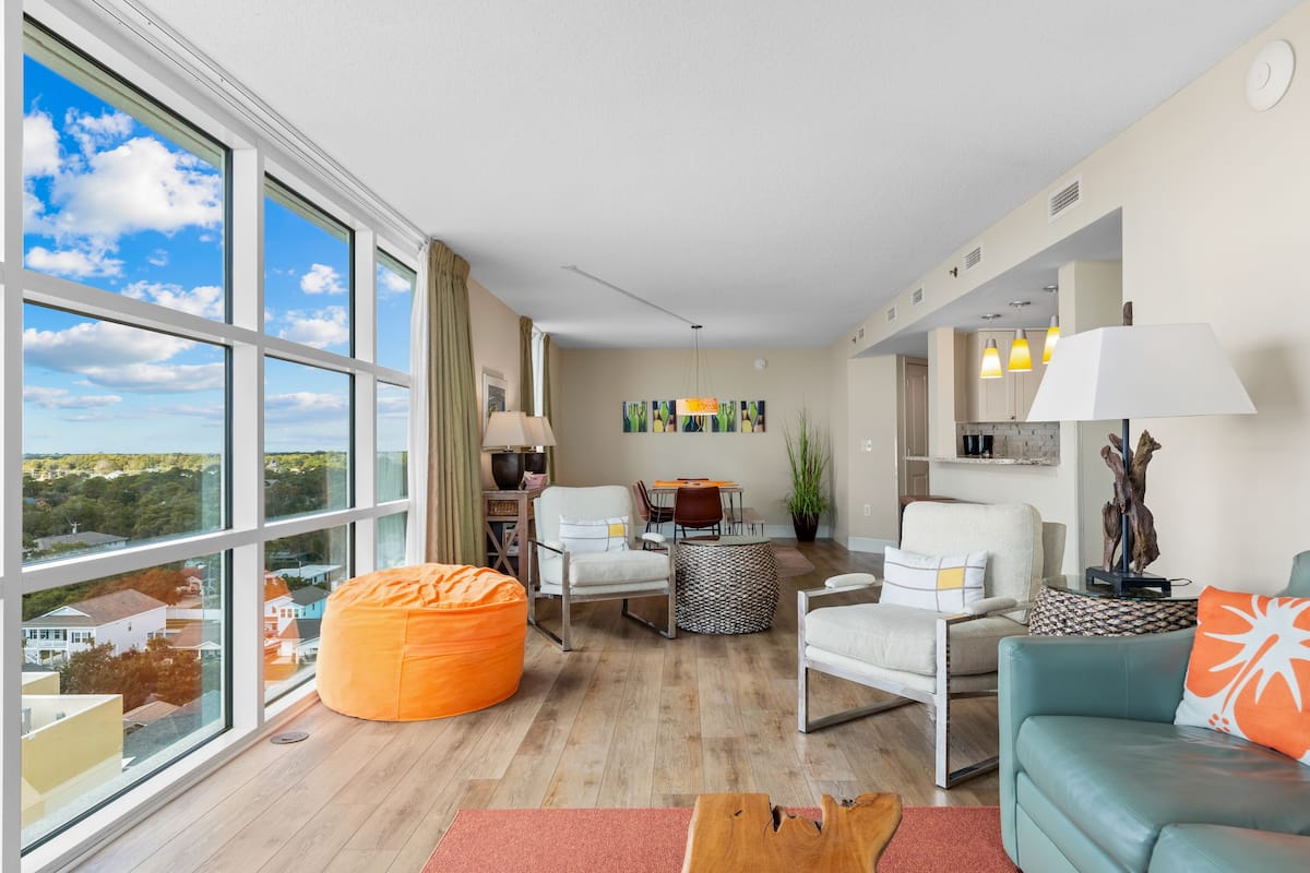 A modern living room with large windows, a dining area, and a kitchen.