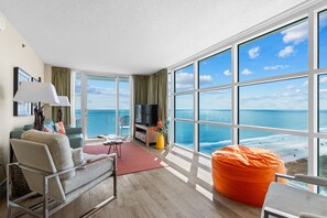 Una sala de estar moderna con un ventanal grande que da al mar, un sofá cómodo, un televisor y un sillón de peluche naranja.