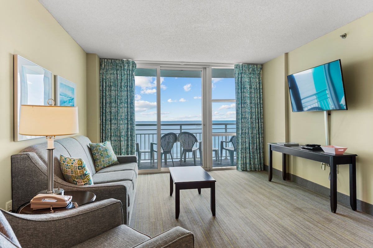 A hotel room with a balcony, a sofa, a coffee table, a TV, and a desk.