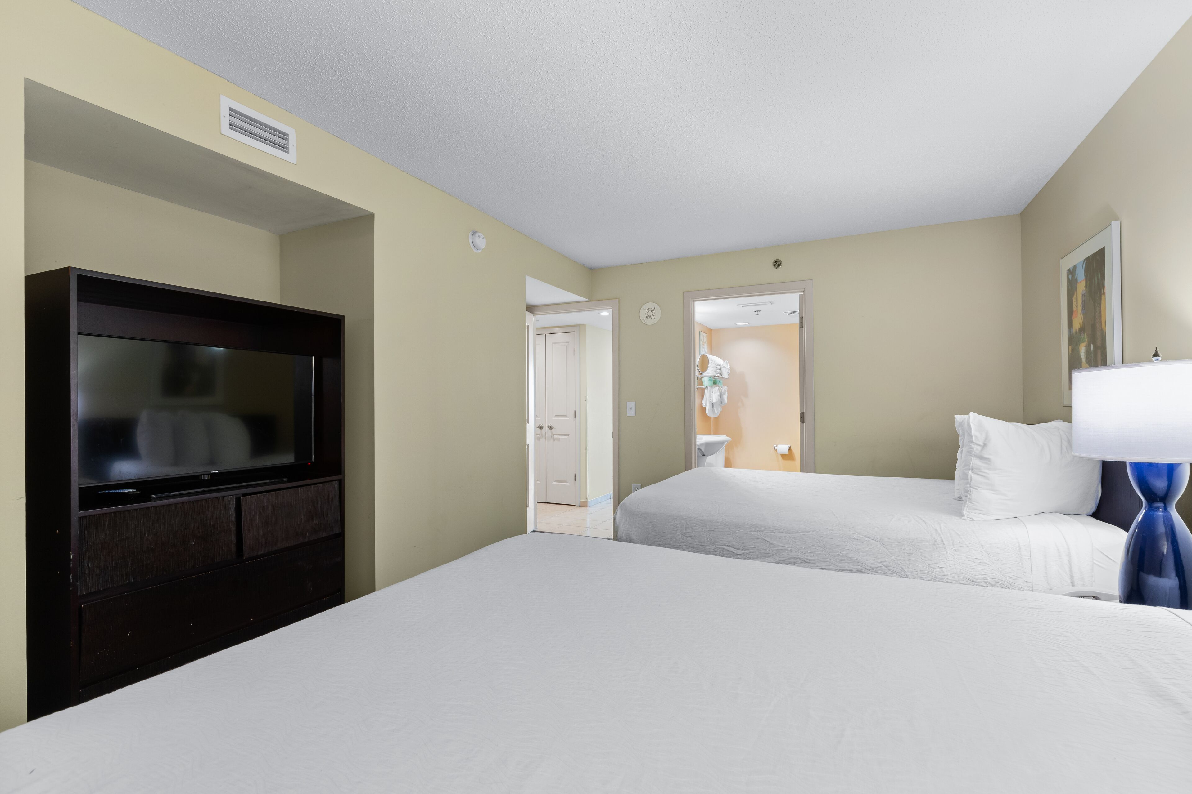 A hotel room with a large flat-screen TV, two beds, a bedside table with a lamp, and a door leading to a bathroom.