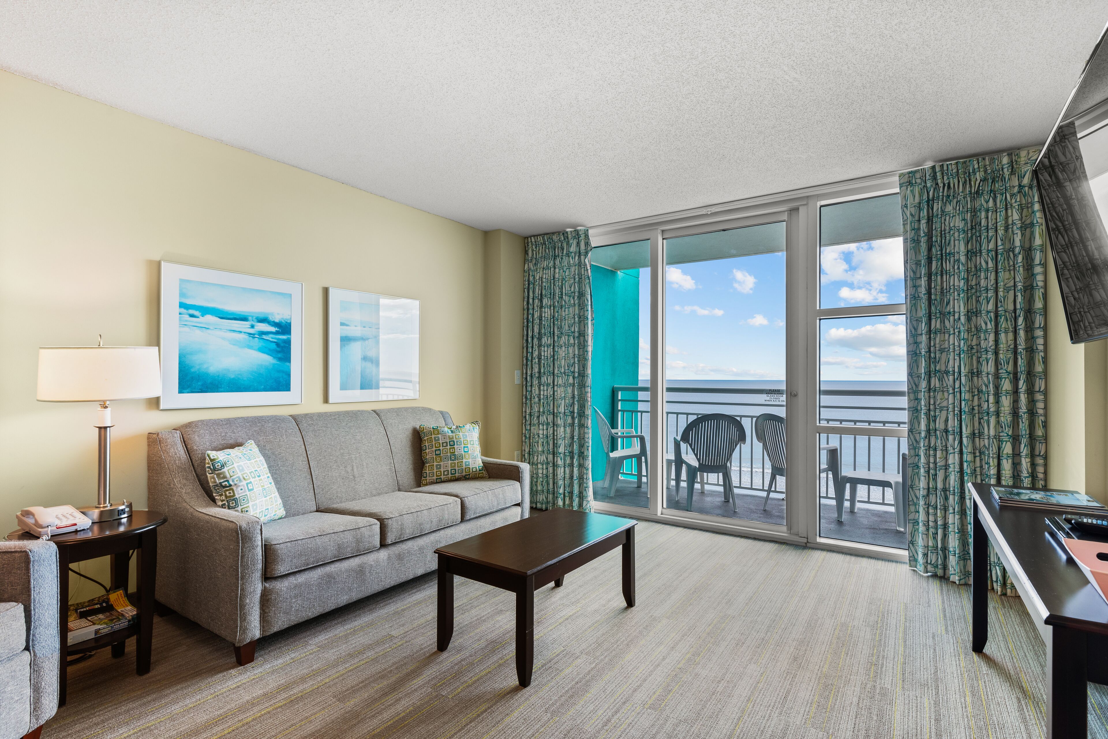 A hotel room with a sofa, a coffee table, a lamp, and a TV. There are two framed pictures on the wall, and a balcony with a view of the ocean.