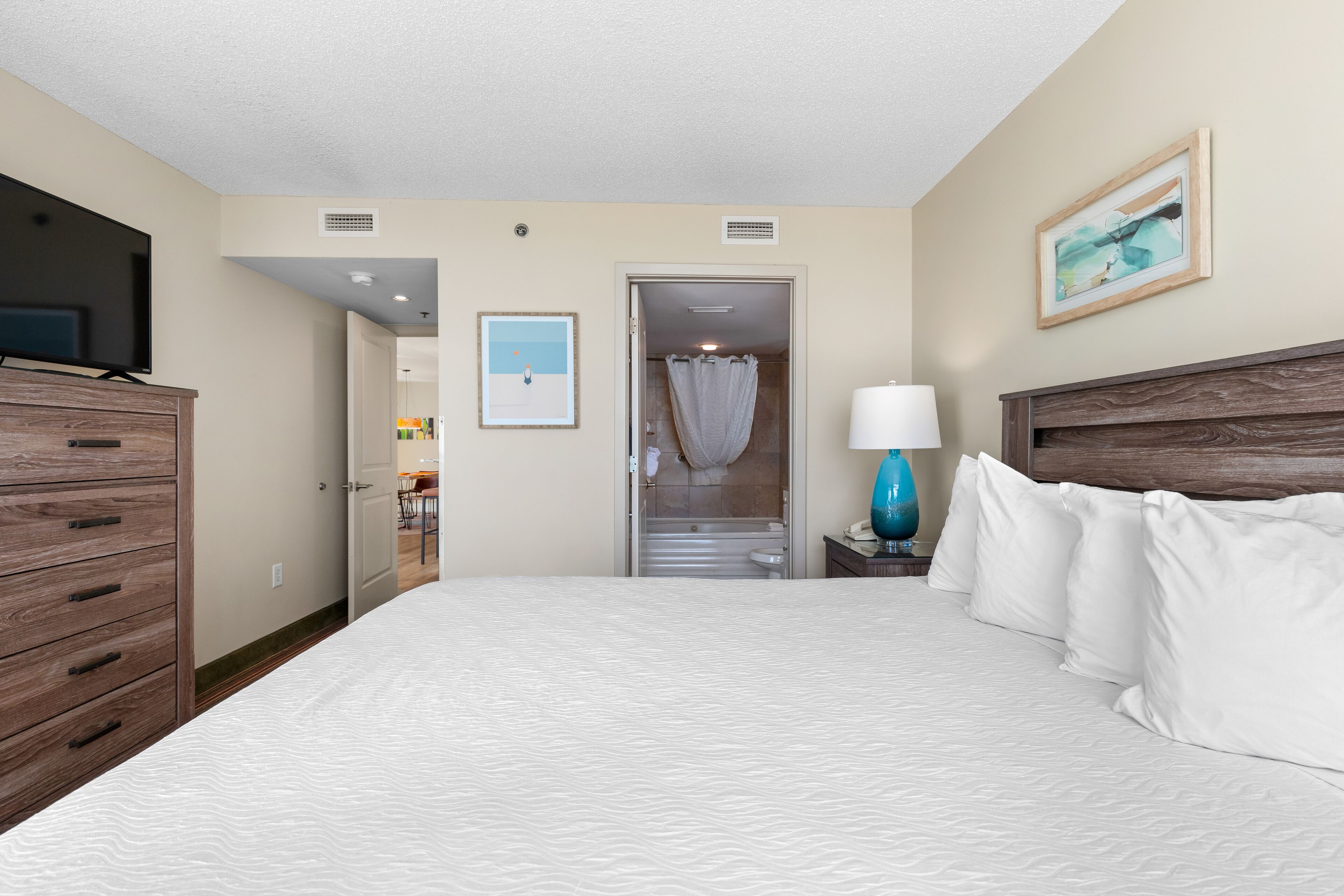 A hotel room with a large bed, a wooden dresser, a television, a bathroom with a shower curtain, and a framed picture on the wall.