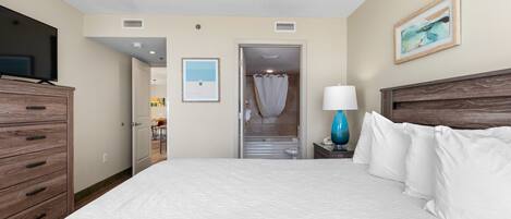 A hotel room with a large bed, a wooden dresser, a television, a bathroom with a shower curtain, and a framed picture on the wall.