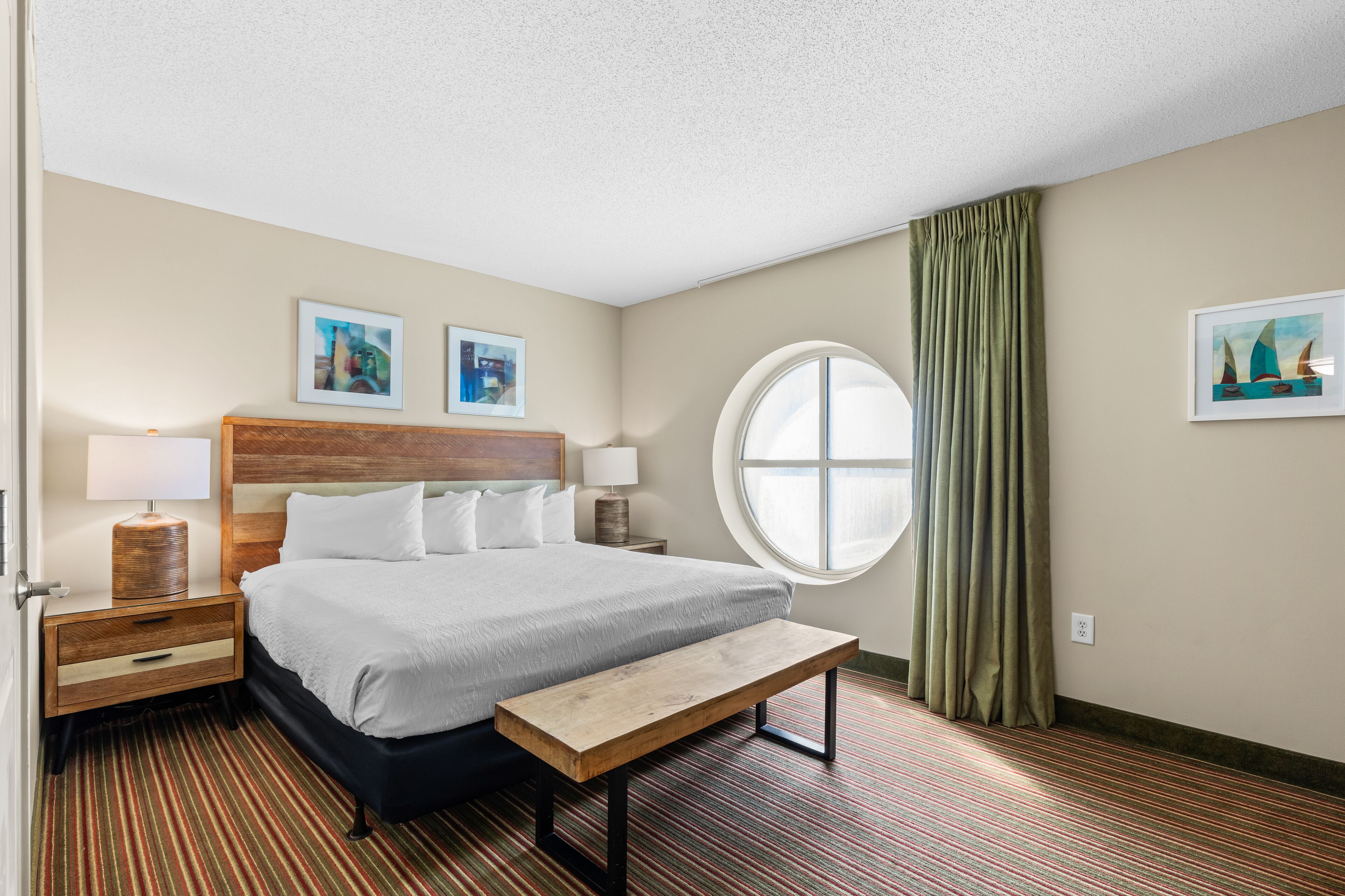 A hotel room with a large bed, a round window, a bench, and two bedside tables with lamps.