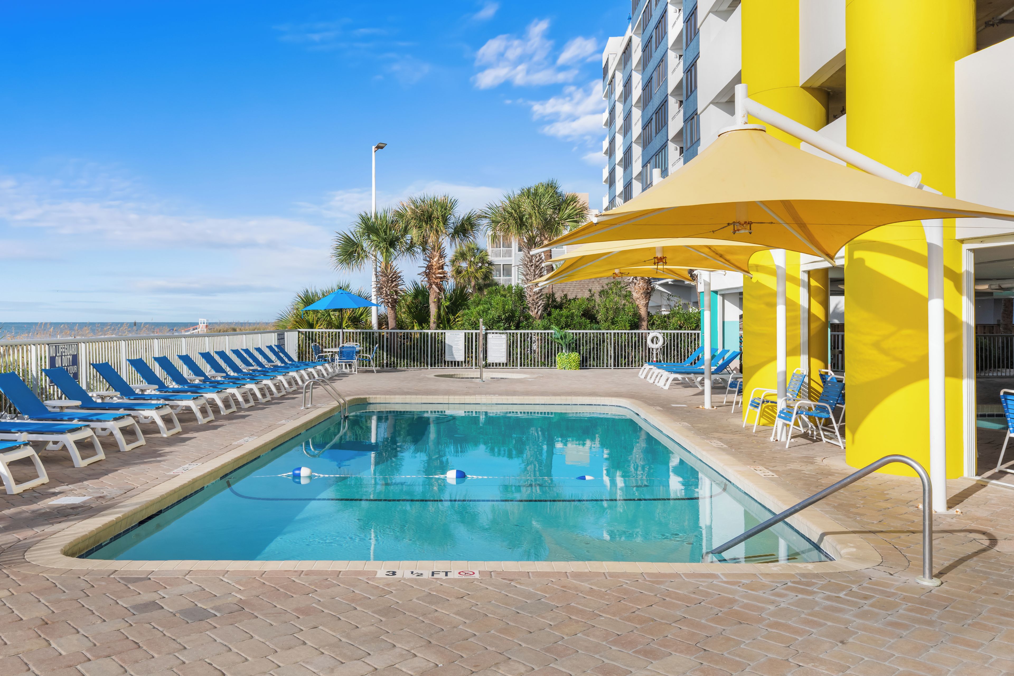 A swimming pool with blue lounge chairs and a yellow umbrella, surrounded by a fence and palm trees.