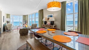 Habitación de hotel moderna con una amplia mesa de vidrio, individuales anaranjados, vista al océano y un rincón de descanso acogedor.
