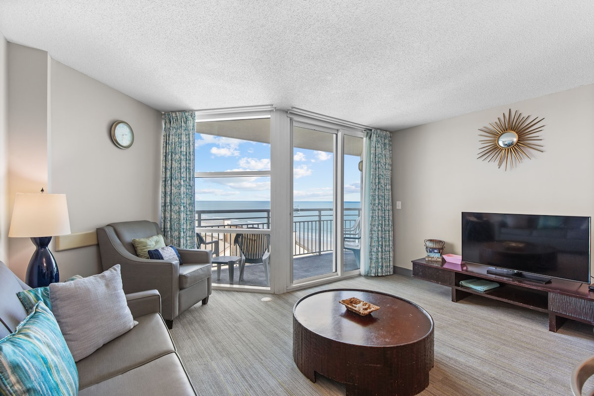 A hotel room with a sofa, a coffee table, a TV, and a balcony with a view of the sea.