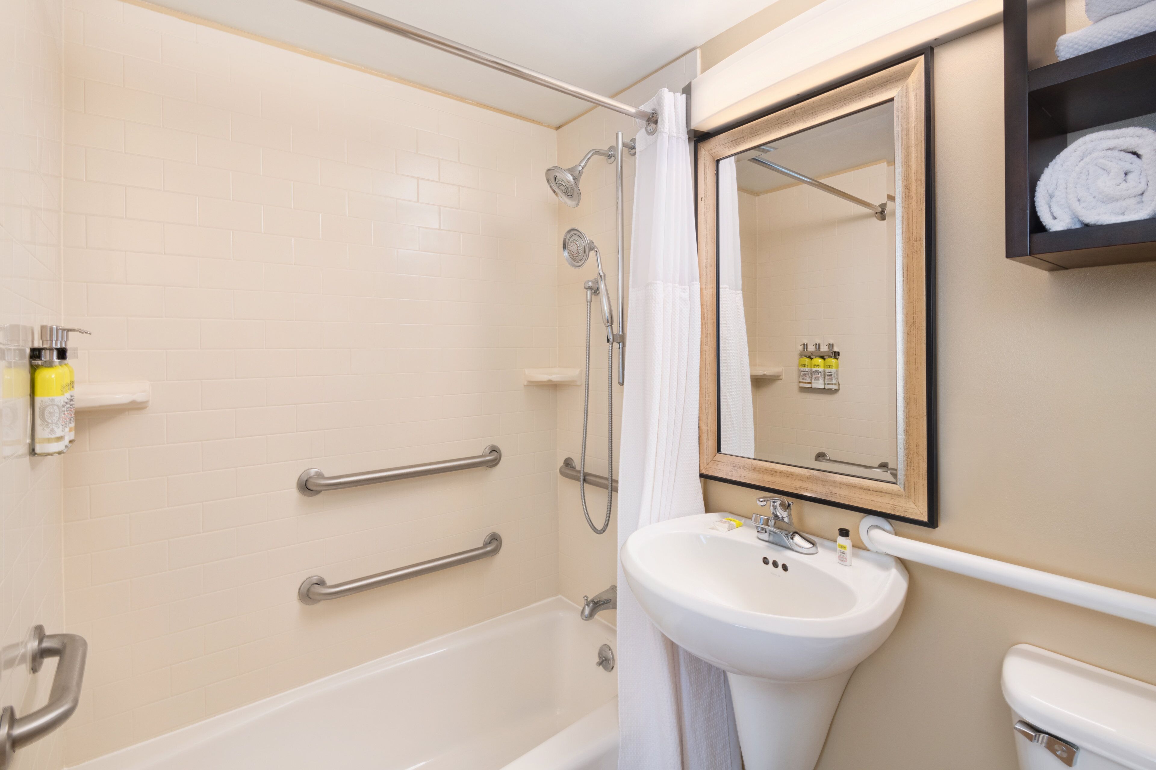A bathroom with a bathtub, a sink, and a mirror. There are handrails and a shower curtain.