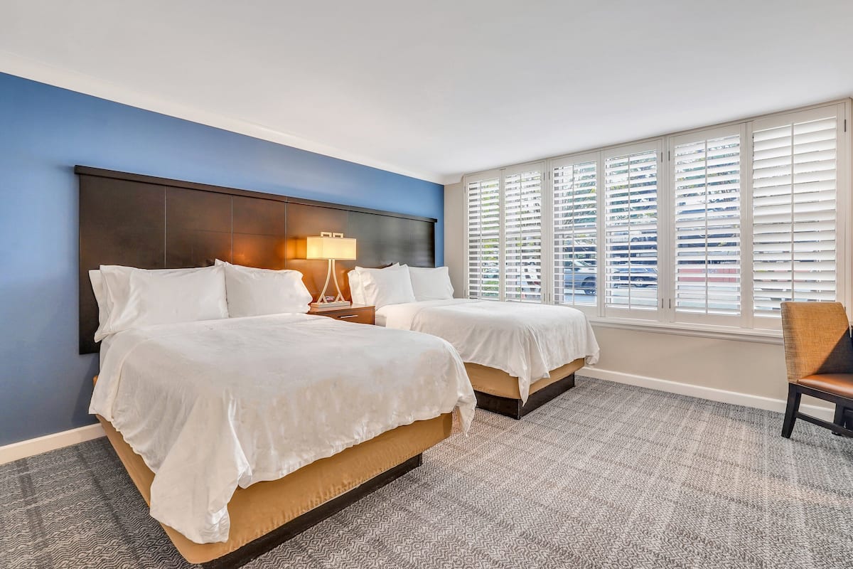 Habitación de hotel con dos camas, una mesita de noche con lámpara, una ventana con persianas y una silla.