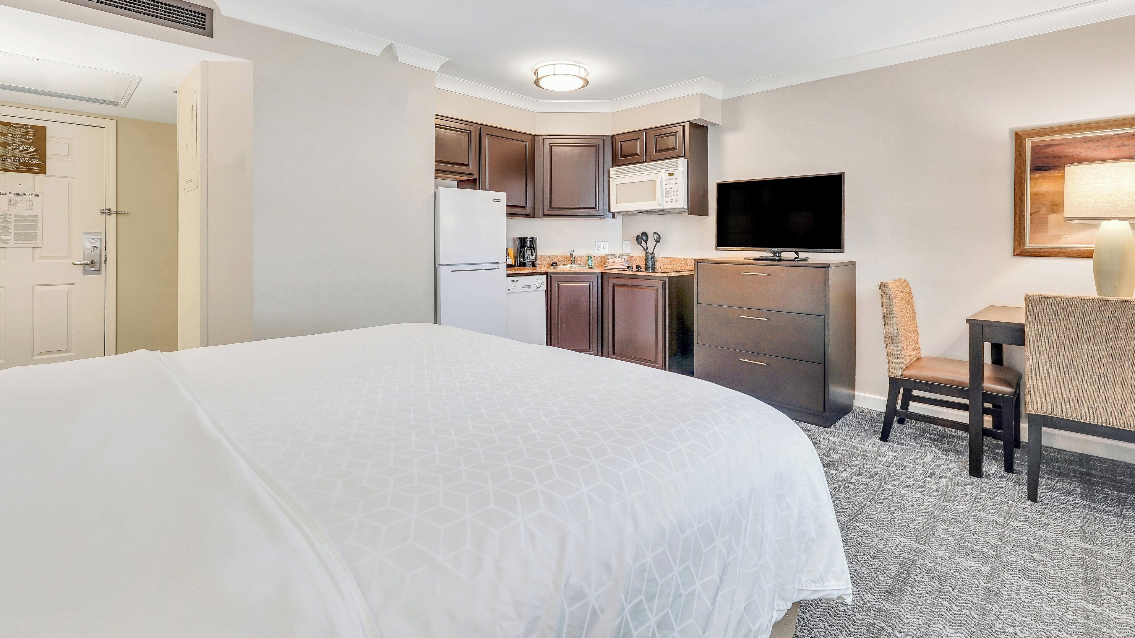 A hotel room with a large bed, a kitchenette with a microwave, refrigerator, and cabinets, a television, and a chair.