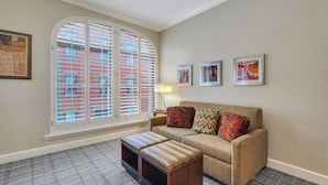 A living room with a beige sofa, two patterned cushions, a small table with a lamp, and a large window with blinds. - Staybridge Suites Savannah Historic District by IHG (Savannah)