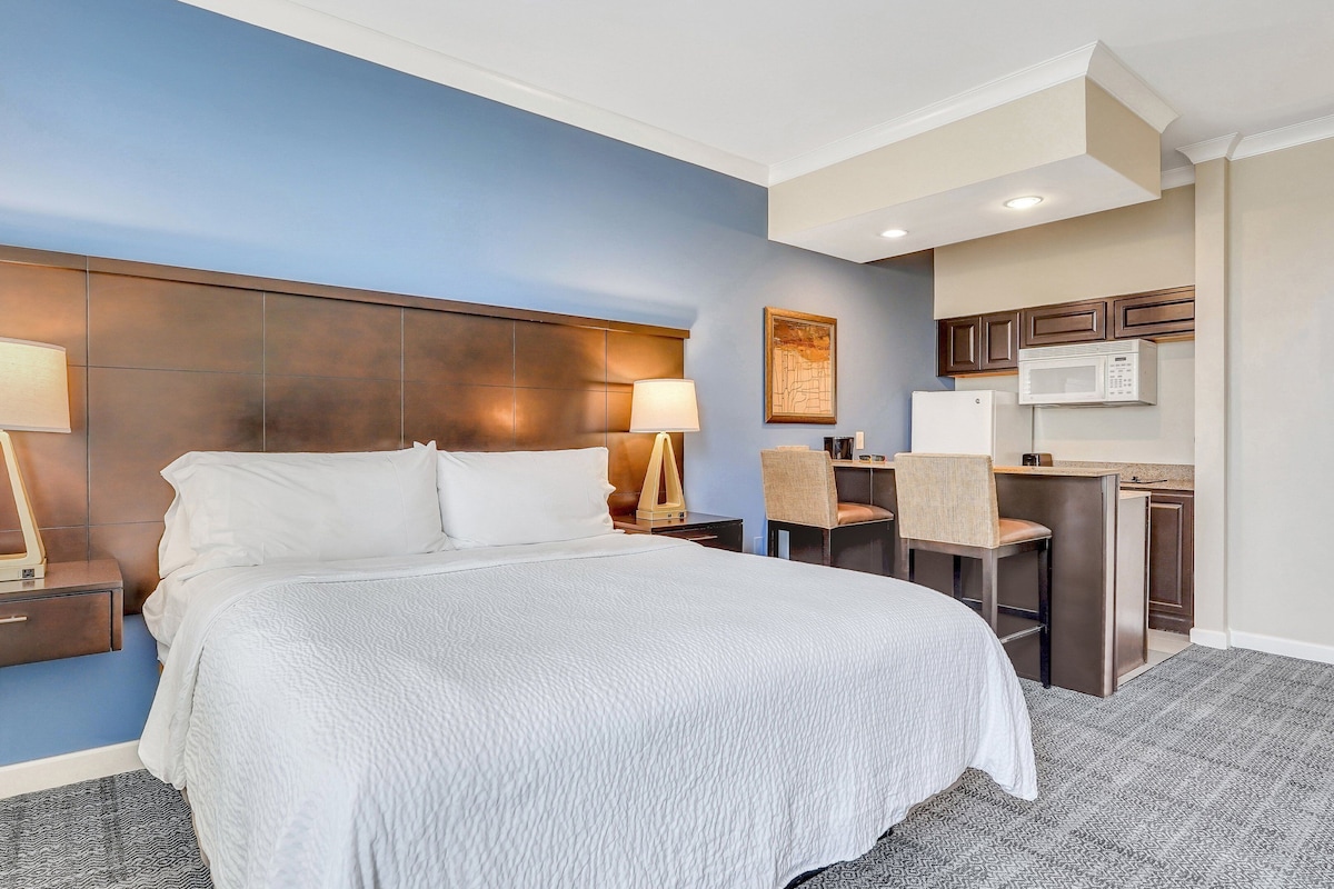 A hotel room with a large bed, two bedside tables with lamps, a small kitchenette, and a blue accent wall.