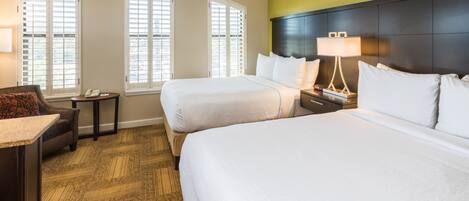 A hotel room with two beds, a desk, a chair, a nightstand, and a lamp.