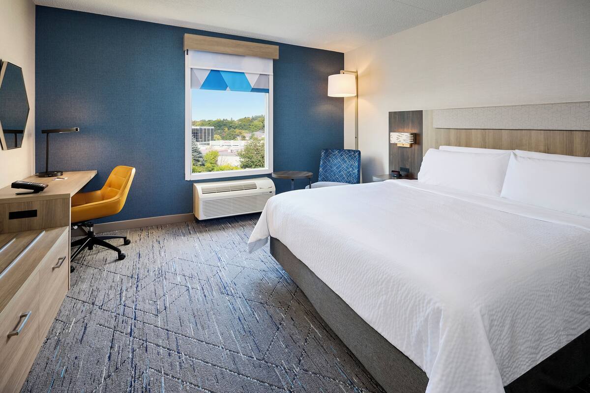 A hotel room with a large bed, a desk with a chair, a window with a view, and a lamp.