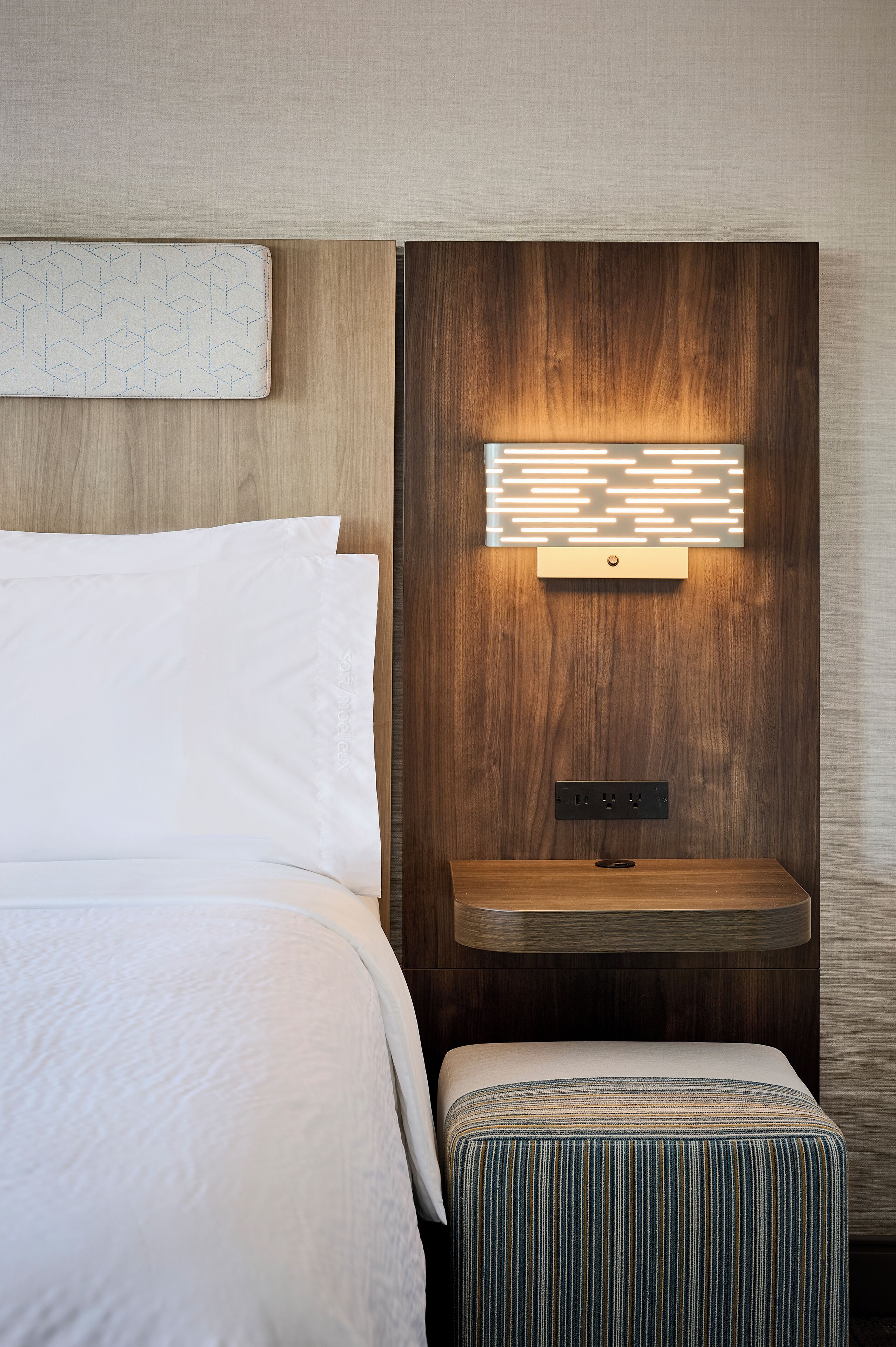 Un lit bien fait, avec du linge de lit blanc, une table de chevet agrémentée d’un coussin à motifs et un luminaire fixé au mur.