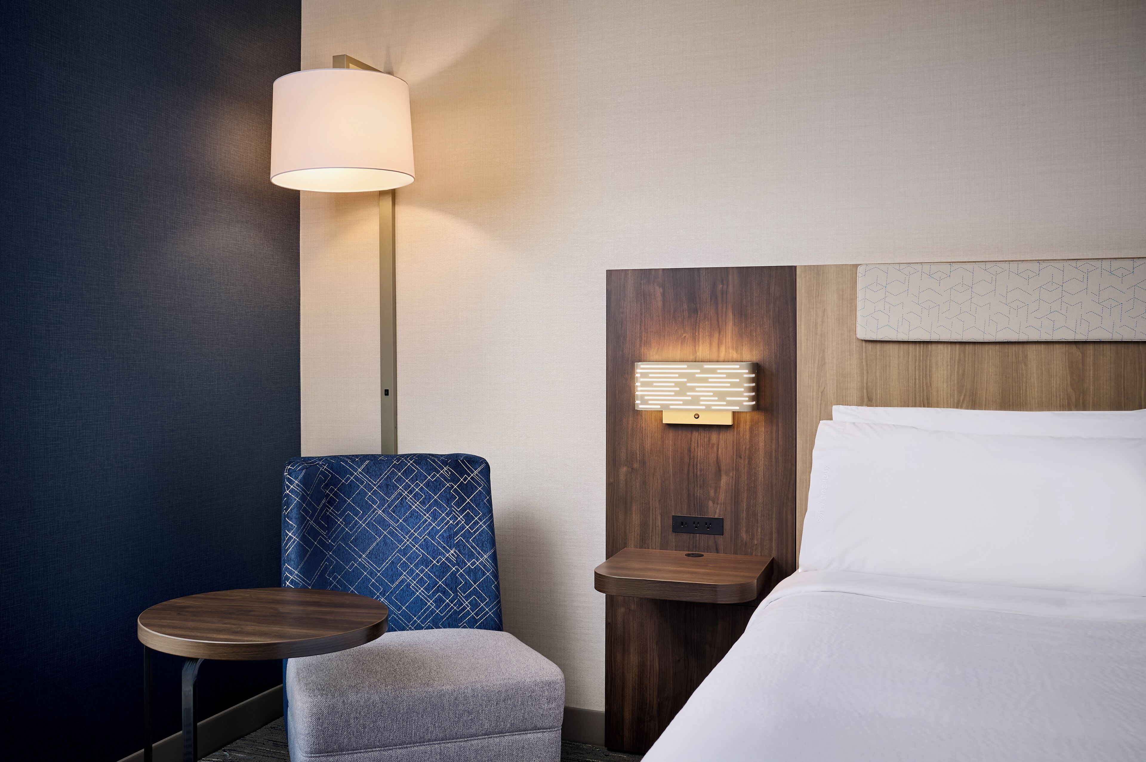 A hotel room with a bed, a nightstand, a lamp, a chair, and a small table.