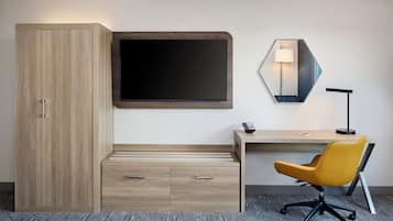 A hotel room with a wooden wardrobe, a flat-screen TV, a desk with a chair, and a wall-mounted mirror.
