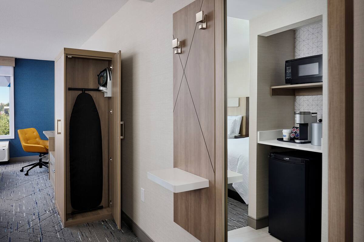 A hotel room with a large mirror, a bed, a small kitchenette, and a seating area with a yellow chair.