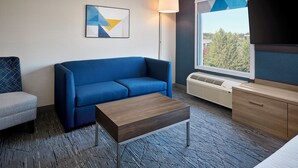 A modern hotel room with a blue sofa, a small wooden table, a grey armchair, a flat-screen TV, and a window with a view of trees. - Holiday Inn Express and Suites Kitchener Southeast by IHG (Kitchener)