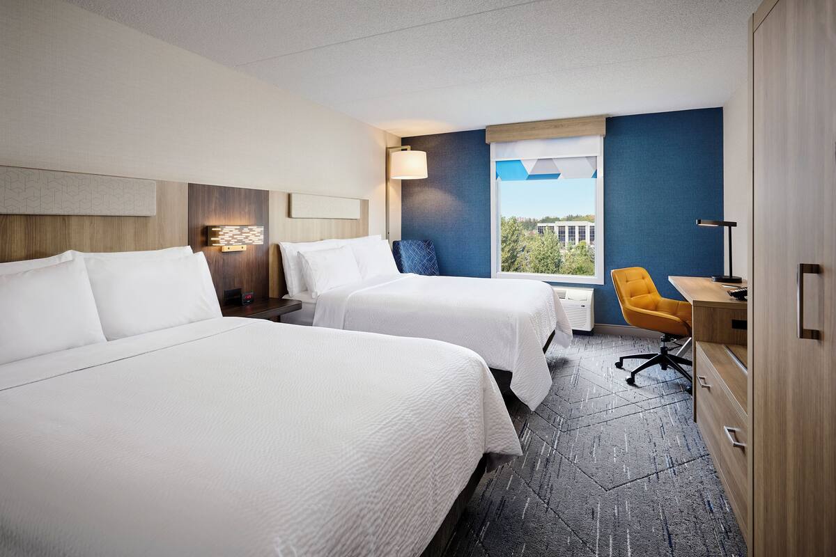 A hotel room with two beds, a desk, a chair, a lamp, and a window with a view.