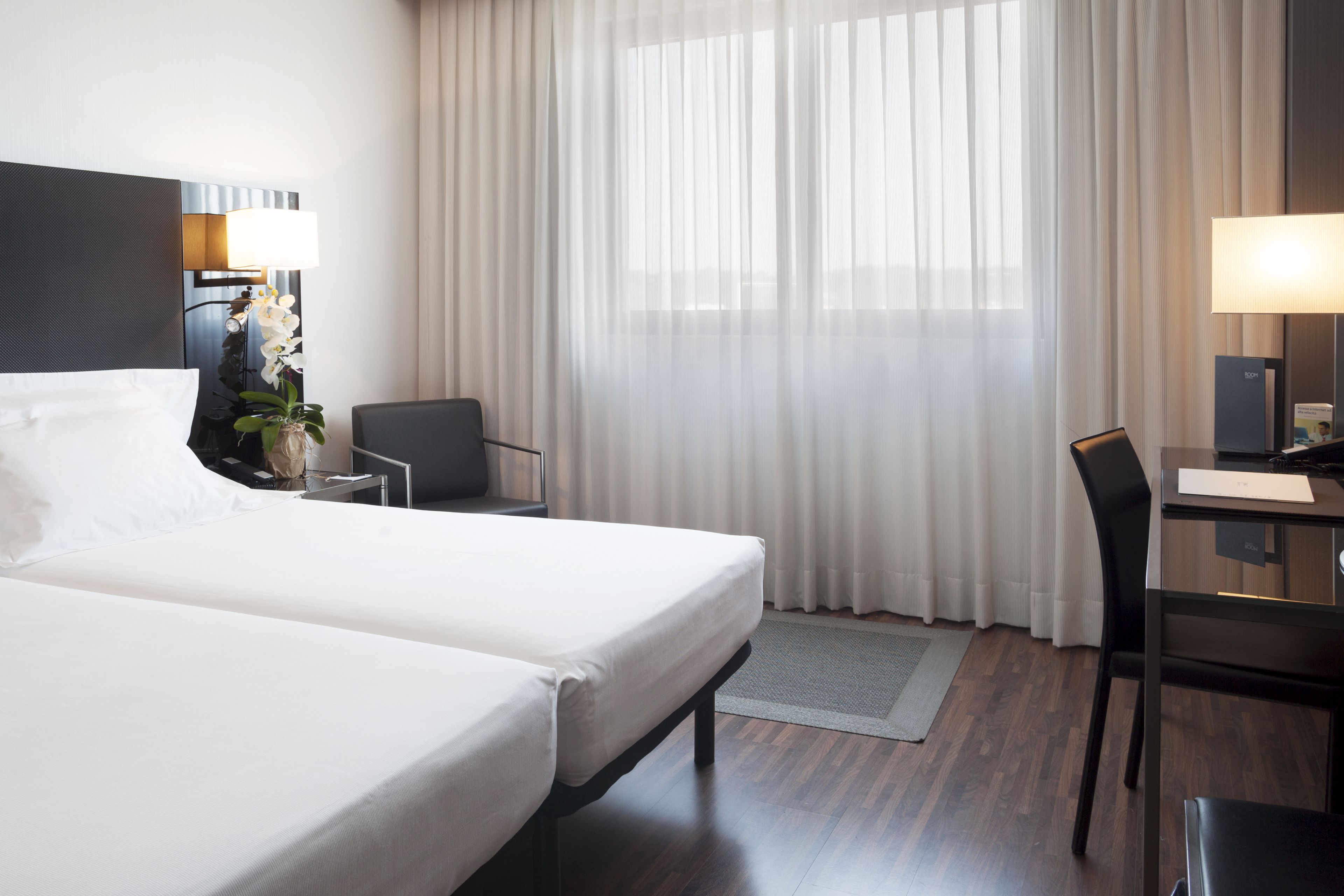 A hotel room with a large bed, a desk, a chair, a lamp, and a window with sheer curtains.