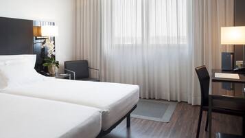 A hotel room with a large bed, a desk, a chair, a lamp, and a window with sheer curtains.