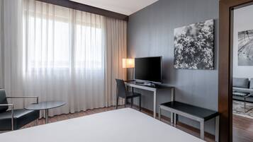 A hotel room with a large bed, a desk with a TV, a chair, a small table, and a sofa.