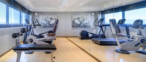 A modern gym with cardio machines, a bench press, and large windows offering a view of the city.