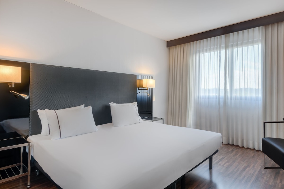 Habitación de hotel con una cama grande, una mesita de noche, una lámpara y un ventanal con cortinas.