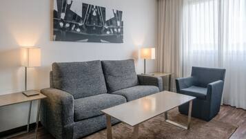 Une chambre d’hôtel moderne avec un canapé gris, un fauteuil assorti, une petite table et deux lampes de chevet. Une grande œuvre d’art abstraite orne le mur.