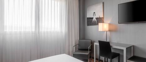 Habitación de hotel moderna con una cama grande, dos sillones, un escritorio, un televisor de pantalla plana, un cuadro en la pared y una ventana con cortinas traslúcidas.