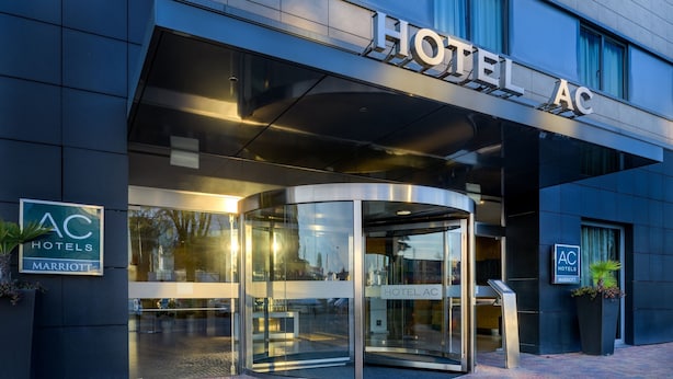 Une entrée d’hôtel moderne avec une porte tournante, une façade en verre et une enseigne indiquant « HOTEL AC » et « AC HOTELS MARRIOTT ».