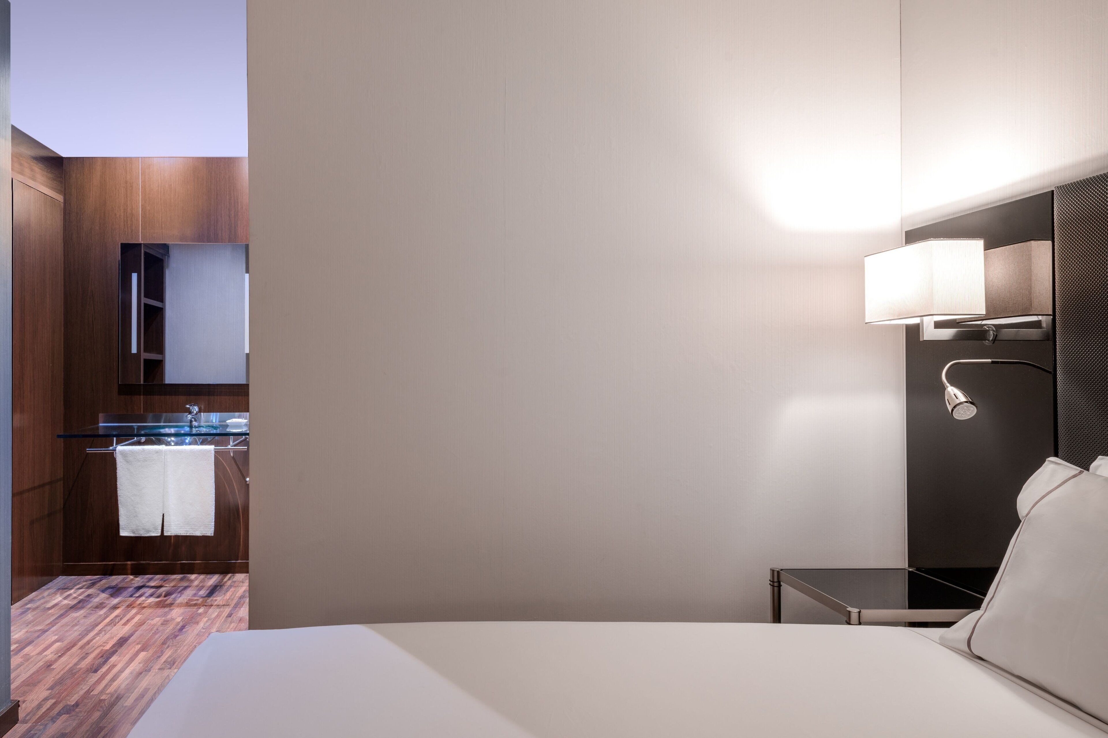 Habitación de hotel con cama, mesita de noche, armario de madera y baño con lavamanos y toallero.