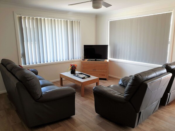 Una sala de estar con un sofá, una mesa de centro con un control remoto, un televisor sobre un mueble de madera y un ventilador de techo.