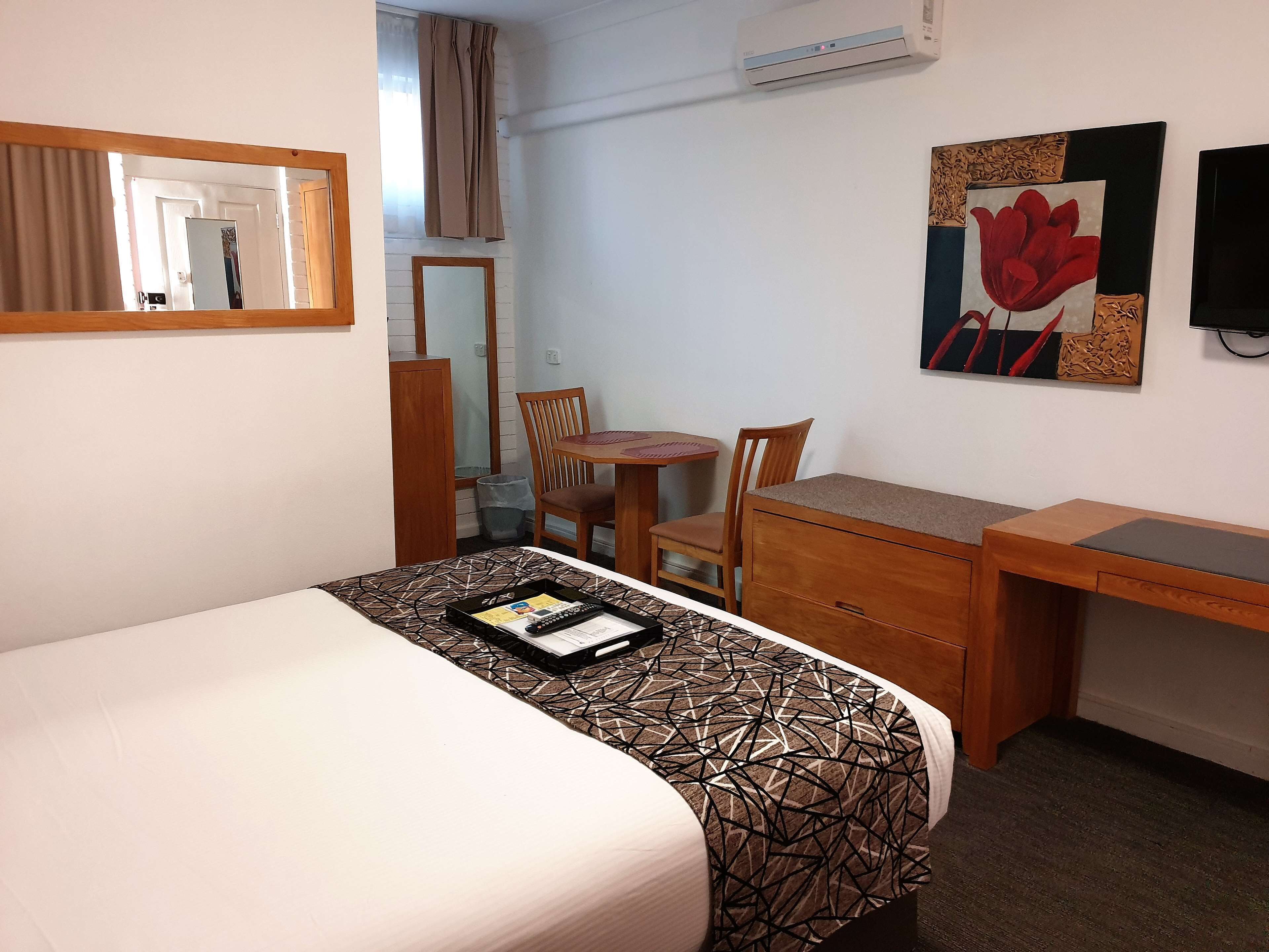 Habitación de hotel con cama, escritorio, silla, mesita pequeña, cesto de basura y aire acondicionado instalado en la pared.