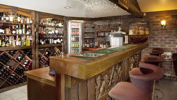 Un coin bar avec des étagères en bois remplies de différentes bouteilles, un réfrigérateur Coca-Cola et une décoration murale sur le thème des navires.