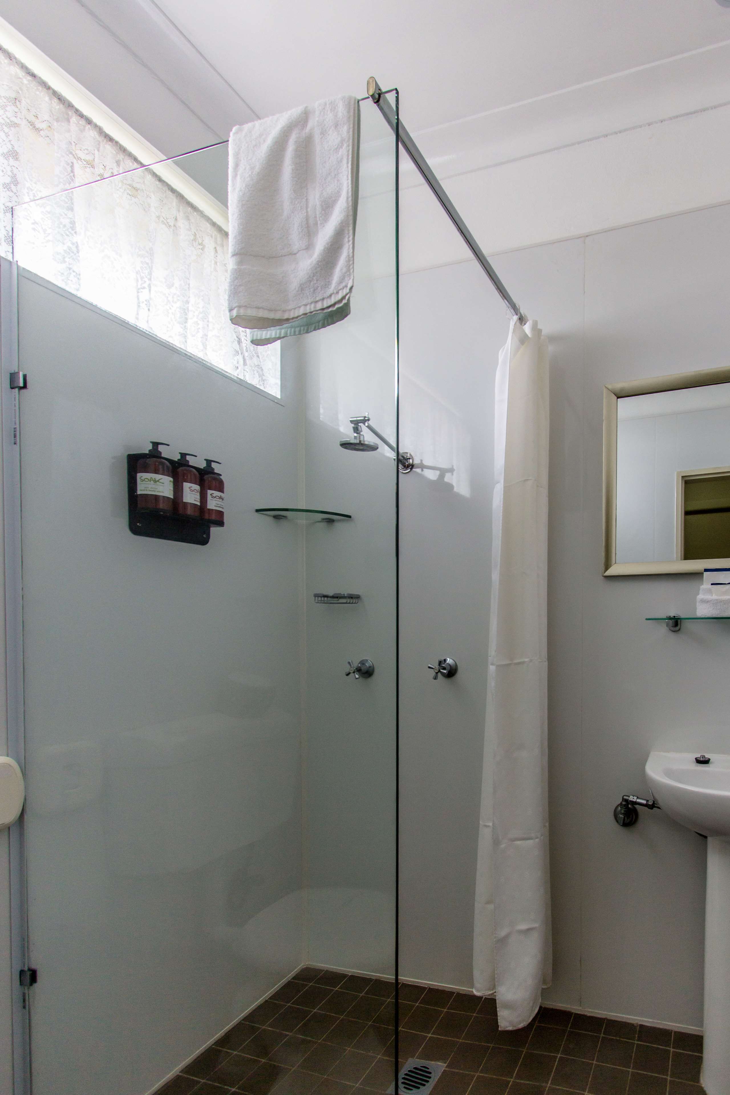 A glass-enclosed shower area with a white towel hanging on a rod, a glass shelf with toiletries, and a sink with a mirror above it.