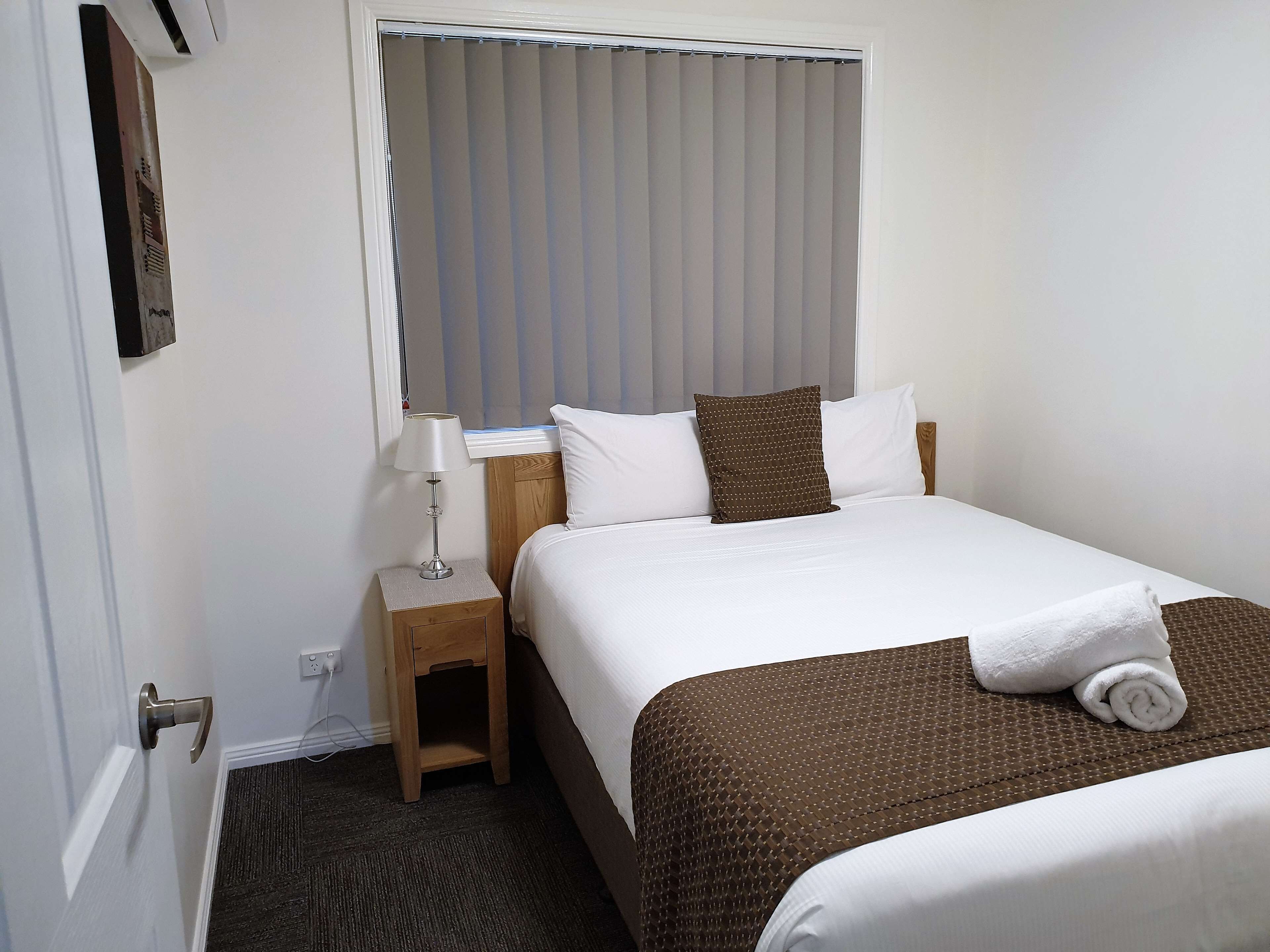 A hotel room with a bed, a nightstand, a lamp, a window with blinds, and a door.