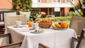 Ett frukostbord dukat med en kopp kaffe, ett glas apelsinjuice, en korg med croissanter, en tallrik med frukt och en liten vas med blommor.
