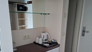 Una kitchenette compacta con microondas, hervidor y cafetera sobre la encimera, un refrigerador pequeño debajo y un espejo arriba.
