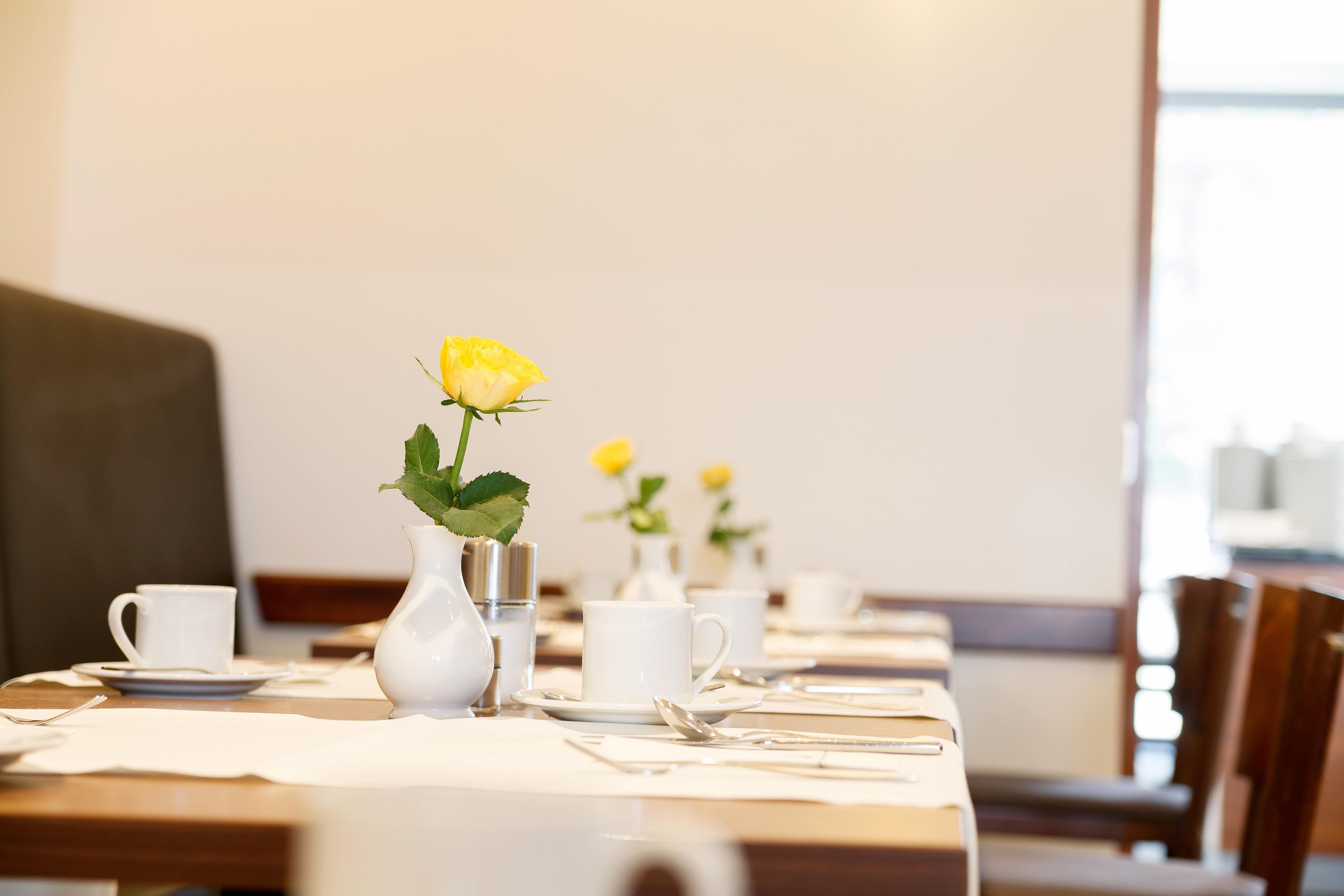 Une table à manger dressée avec des tasses et des soucoupes blanches, ainsi qu’un vase contenant une fleur jaune.