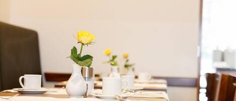 Een eettafel gedekt met witte kopjes, schoteltjes en een vaas met een gele bloem.