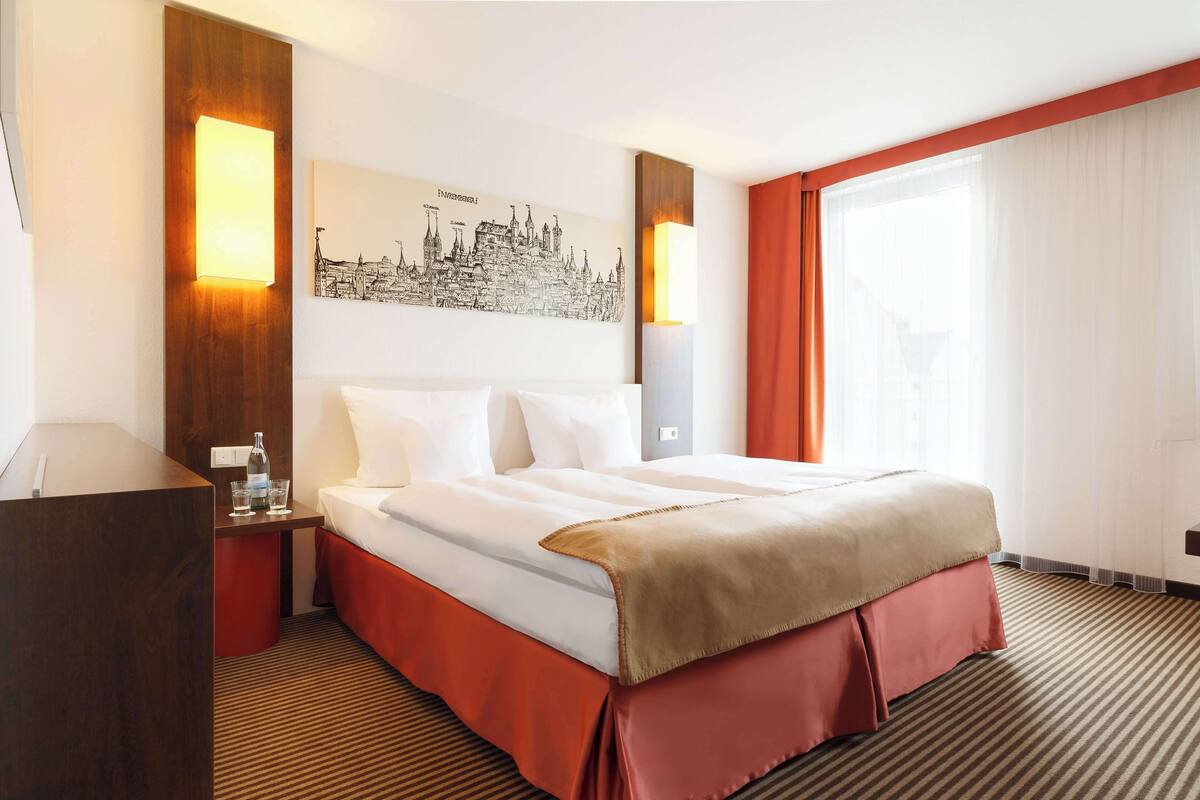 A hotel room with a large bed, a nightstand, a wall-mounted lamp, and a cityscape artwork.