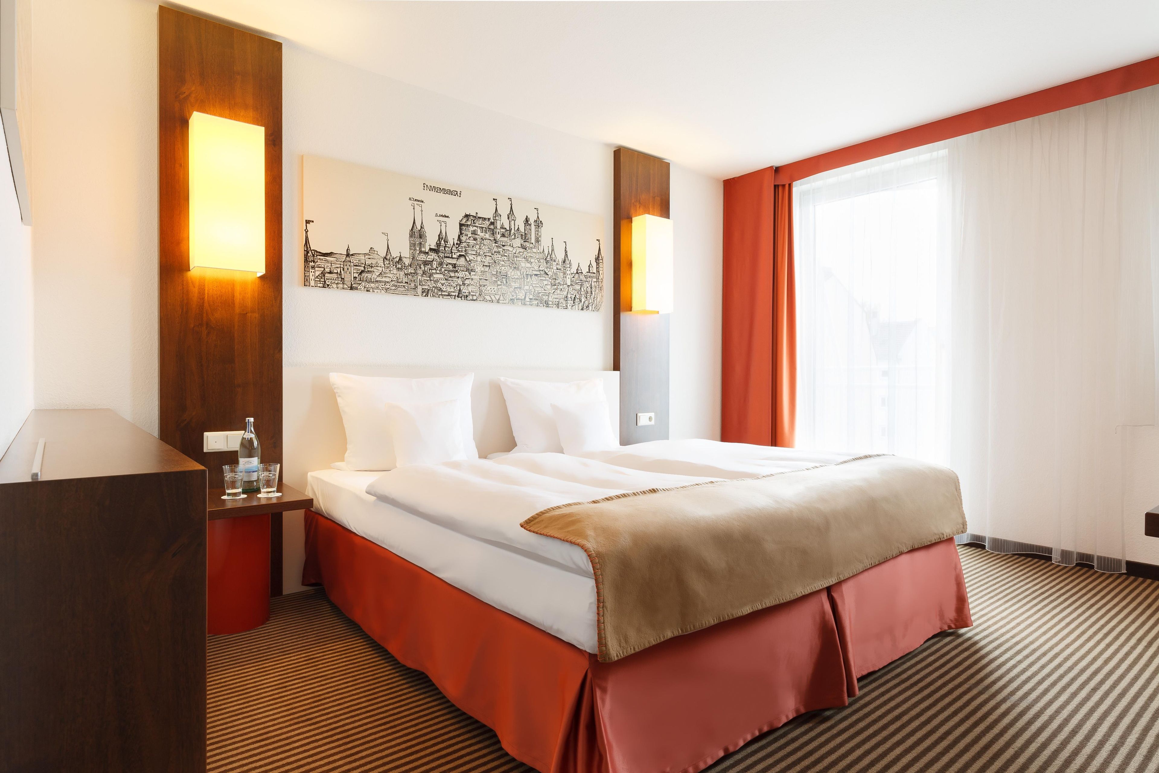 Una camera d'albergo con un letto, un comodino, una bottiglia, bicchieri, un'opera d'arte con una veduta urbana e una finestra con tende.