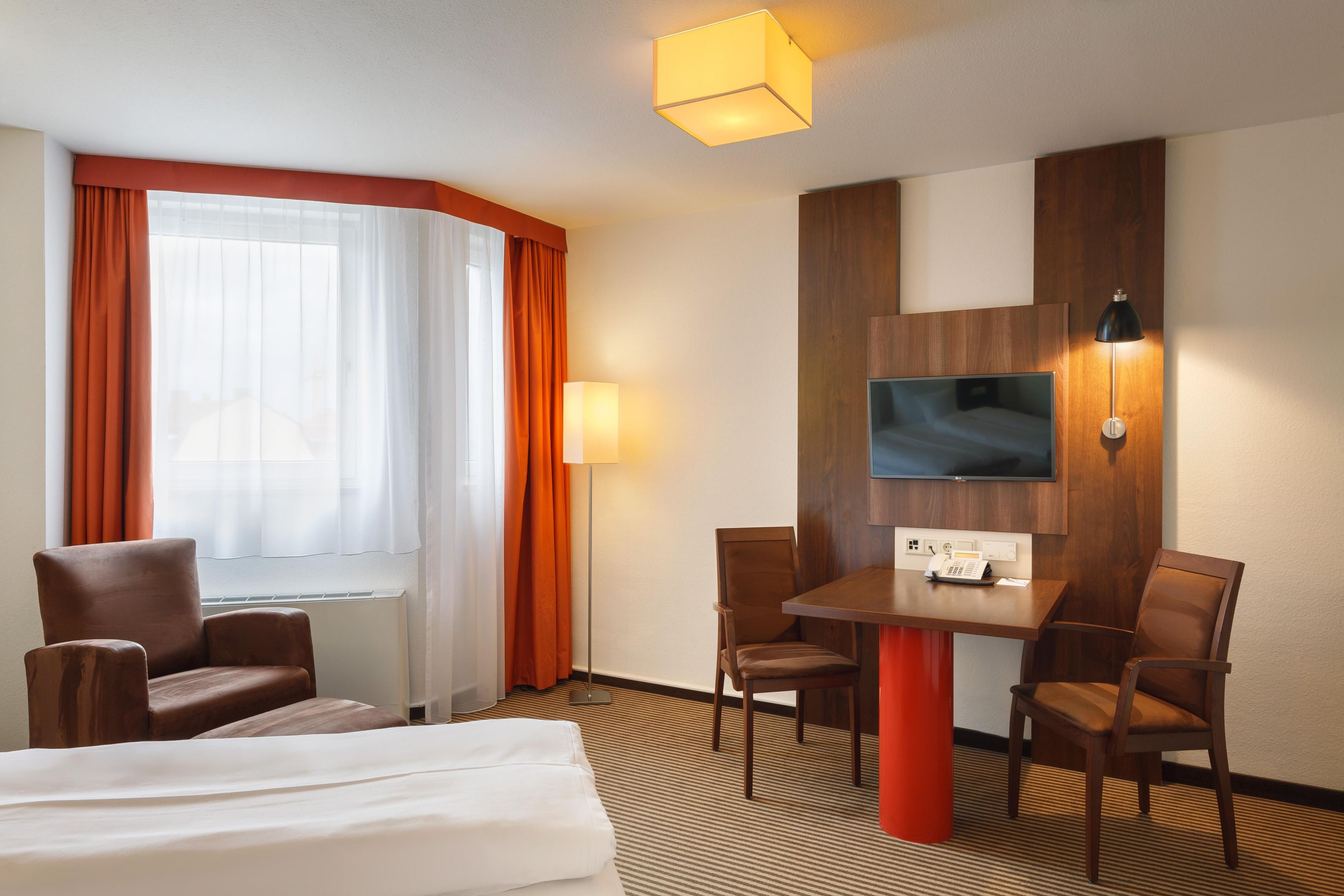 Habitación de hotel con cama, dos sillones, una mesa, televisor de pantalla plana montado en la pared y ventana con cortinas.