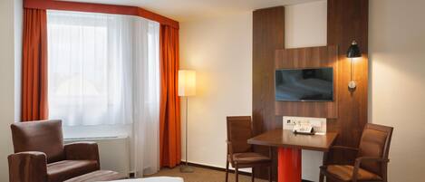 A hotel room with a bed, two armchairs, a table, a flat-screen TV mounted on the wall, and a window with curtains.