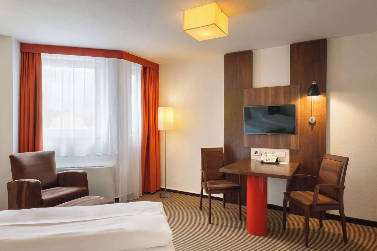 A hotel room with a bed, two armchairs, a dining table, a TV mounted on the wall, and a window with curtains.