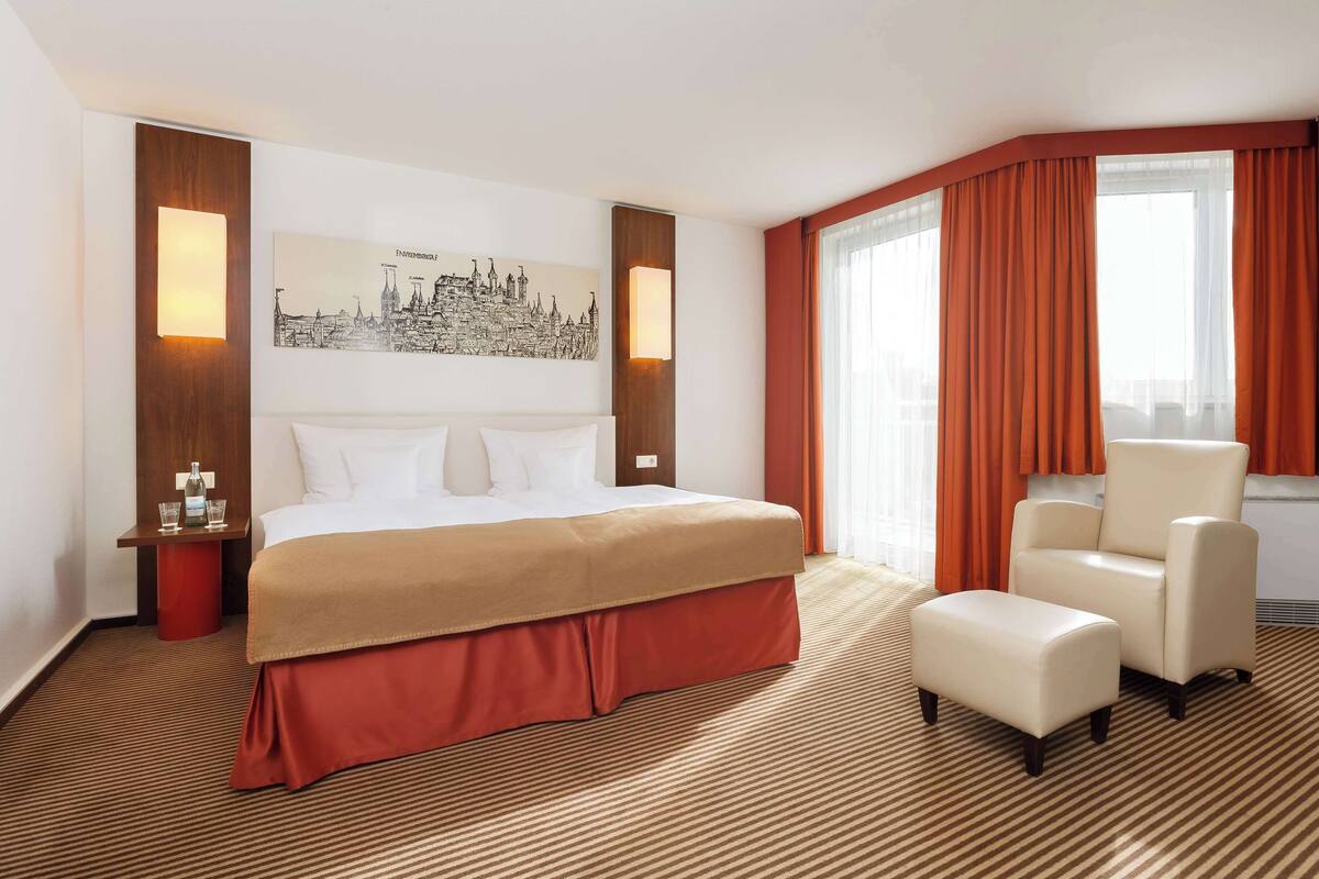 A hotel room with a large bed, a chair, a small table with glasses, and a cityscape painting on the wall.