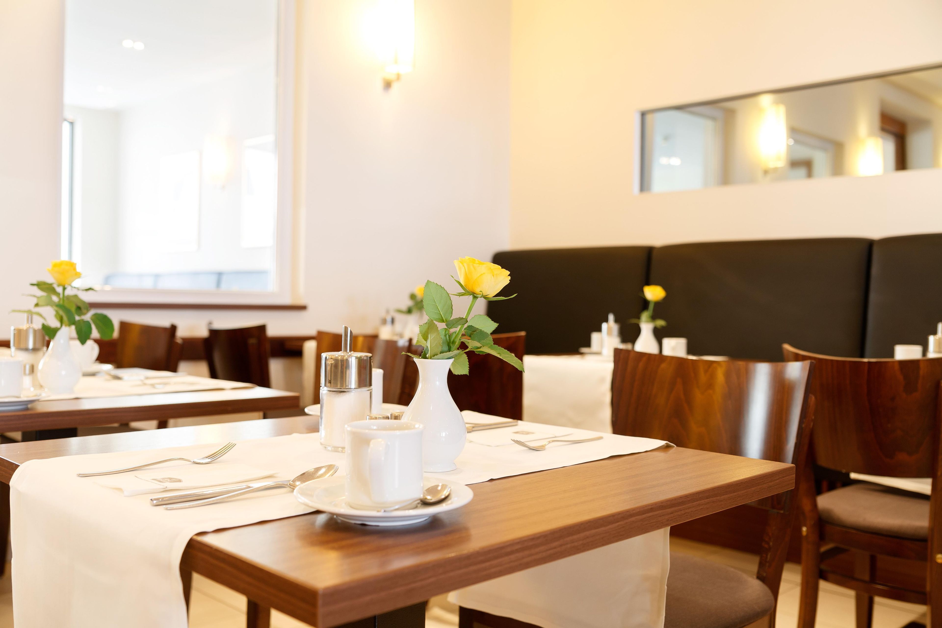 A hotel dining area with tables set for two, each with a white tablecloth, a vase with a yellow flower, and a silver condiment container.
